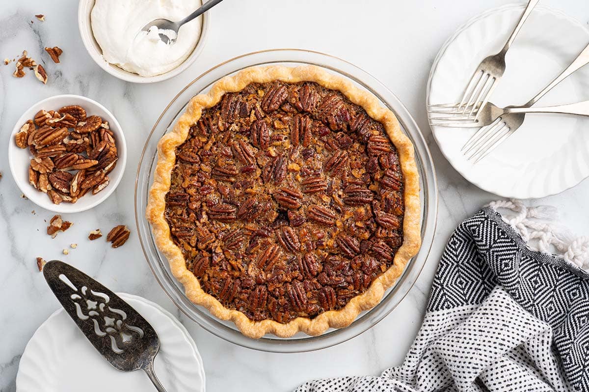 Overhead view of baked pecan pie surrounded by whipped cream, pecans, and serving plates.