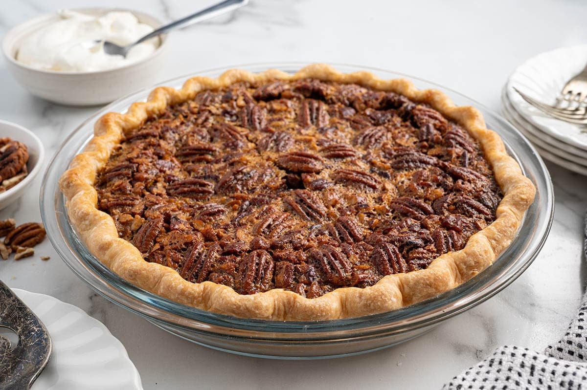 Whole pecan pie with golden crust and glossy pecan topping in a glass pie dish, served with whipped cream.