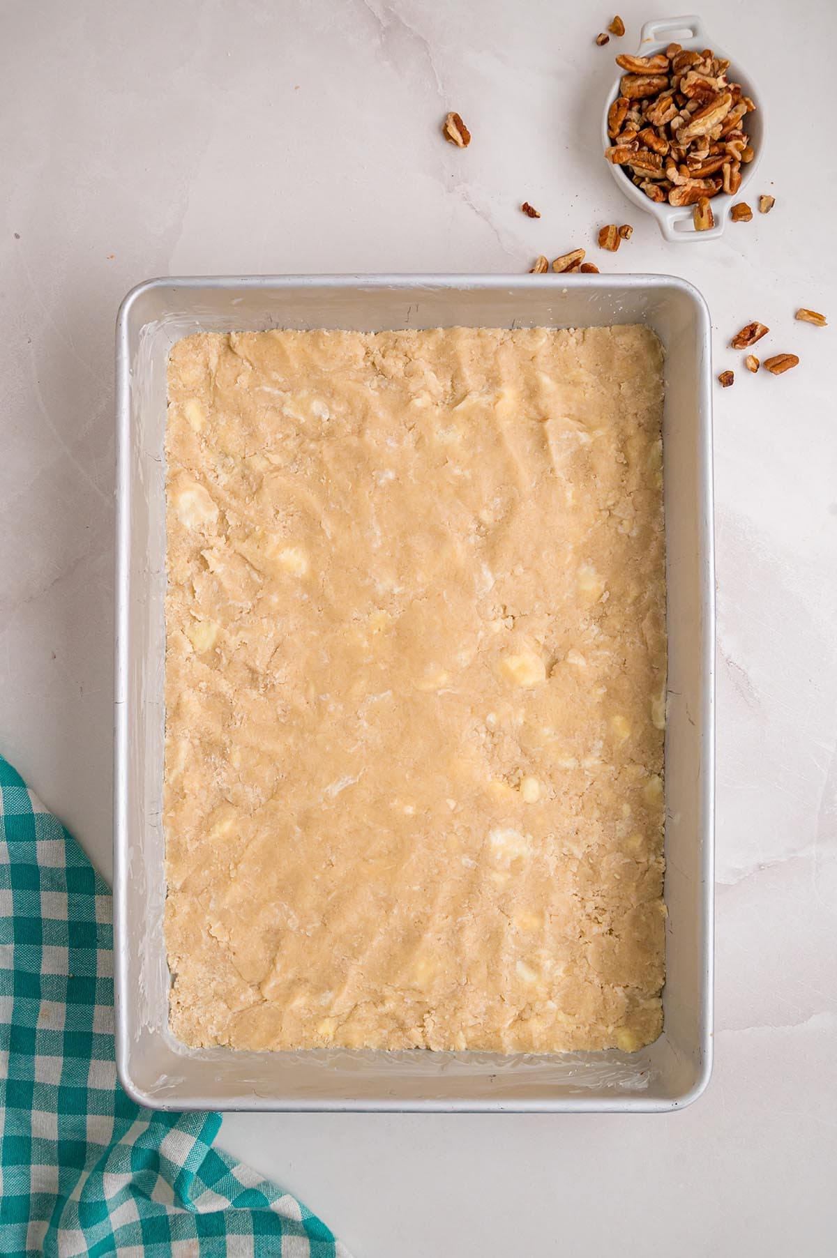 Pecan pie bar crust pressed evenly into a baking pan, with chopped pecans nearby.