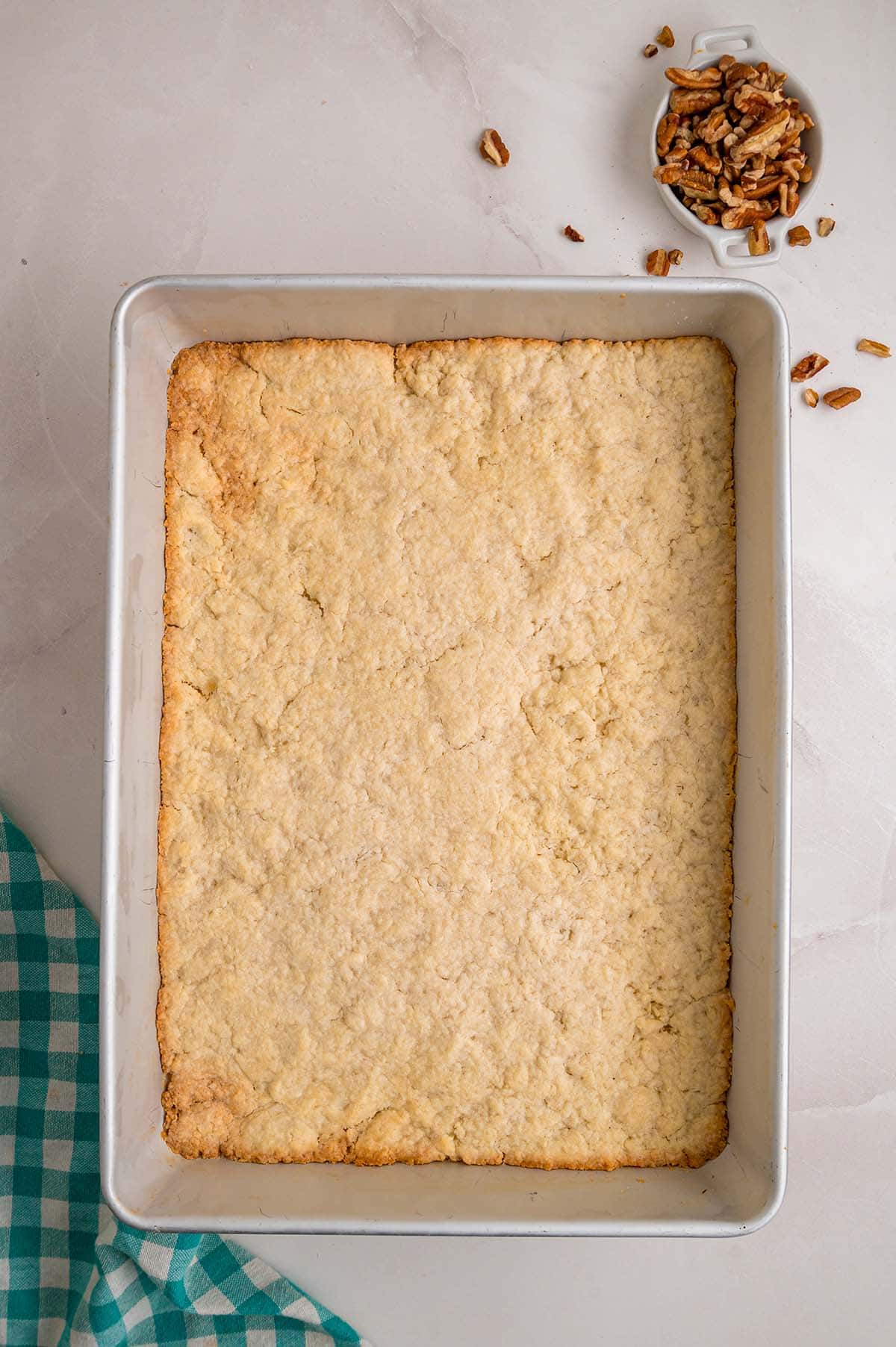 Baked pecan pie bar crust in a rectangular pan, with chopped pecans nearby.
