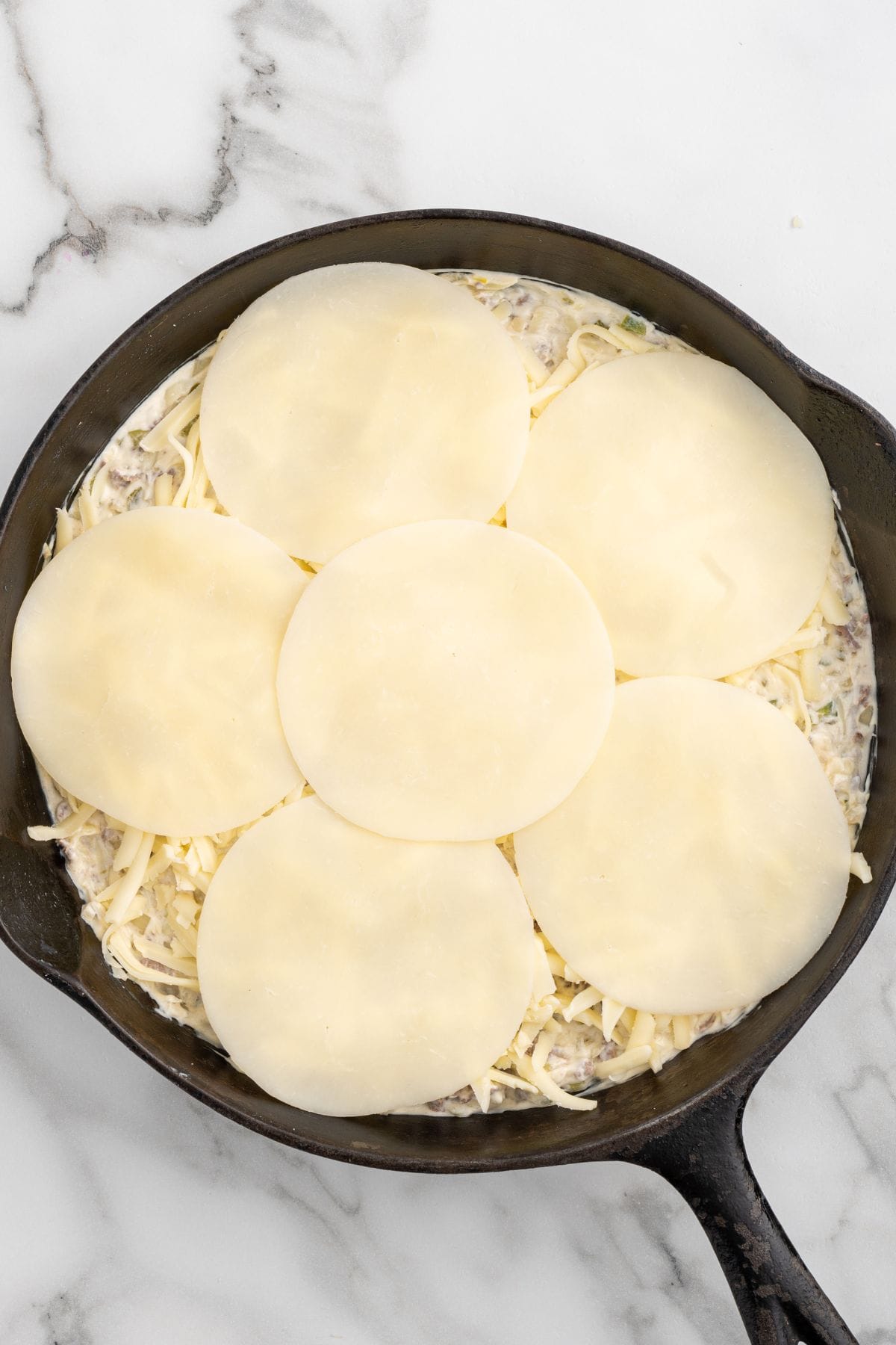 Cheese slices arranged over cheesesteak dip in a cast iron skillet before baking.