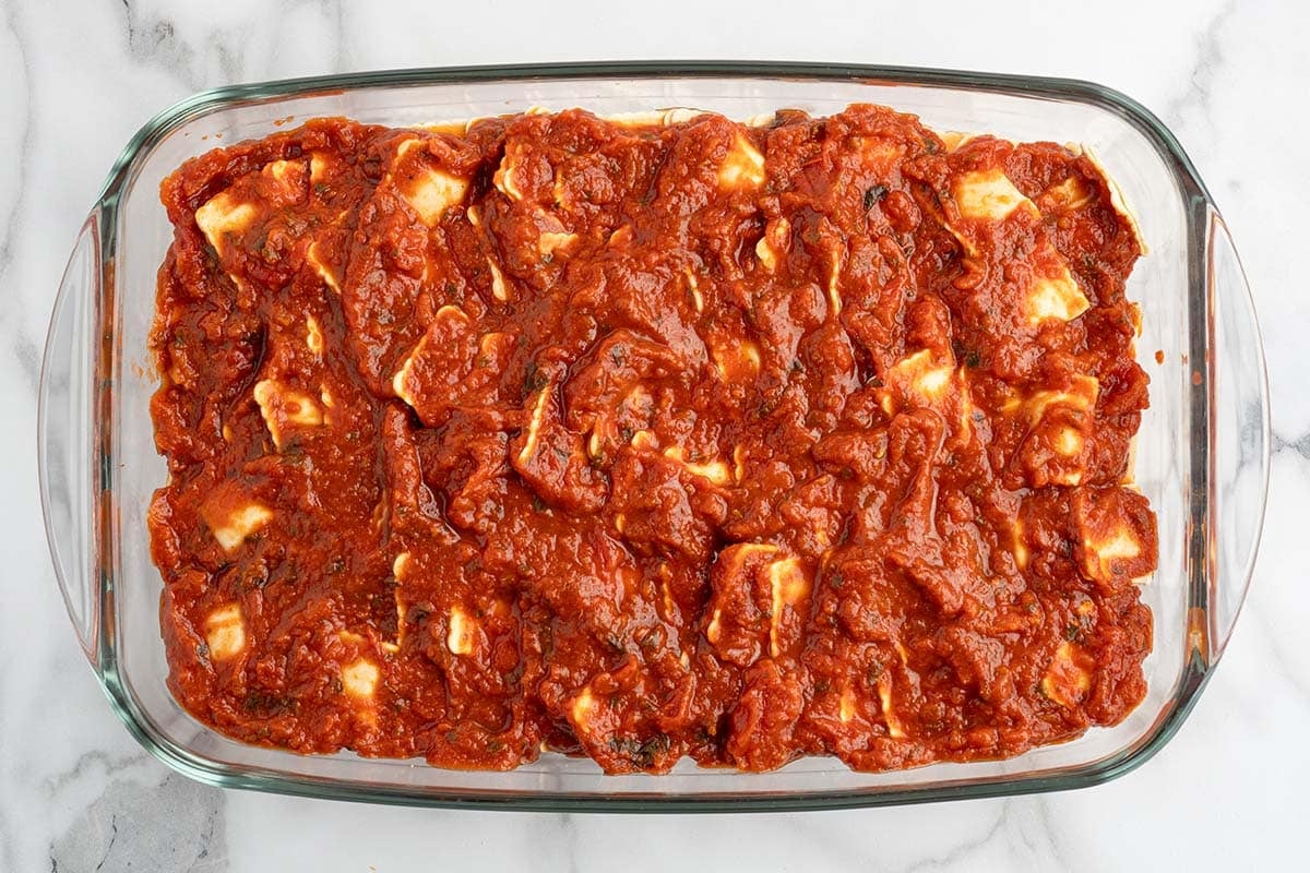 Marinara sauce covering a layer of ravioli in a glass baking dish.