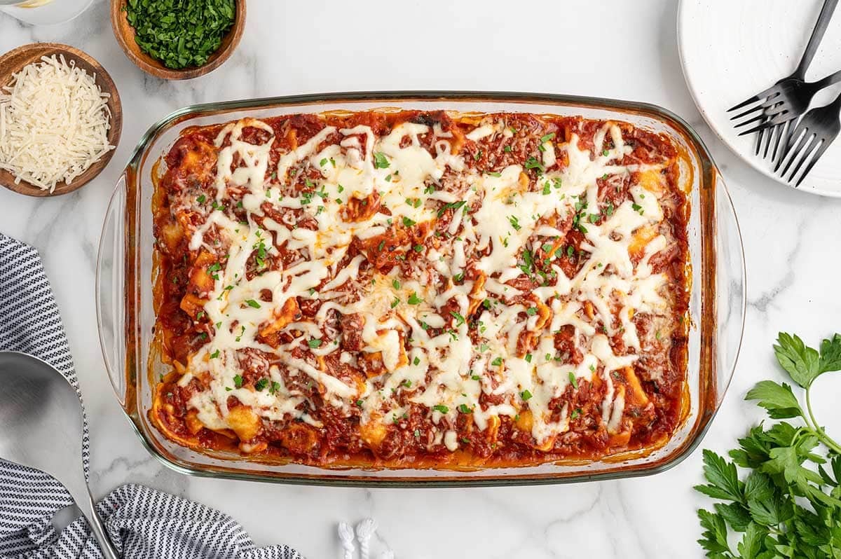 Overhead view of baked ravioli lasagna garnished with parsley, with parsley and cheese bowls nearby.