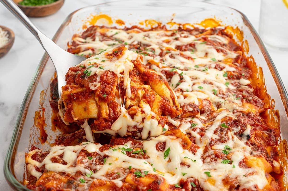 Serving spoon lifting cheesy baked ravioli lasagna from a glass dish.