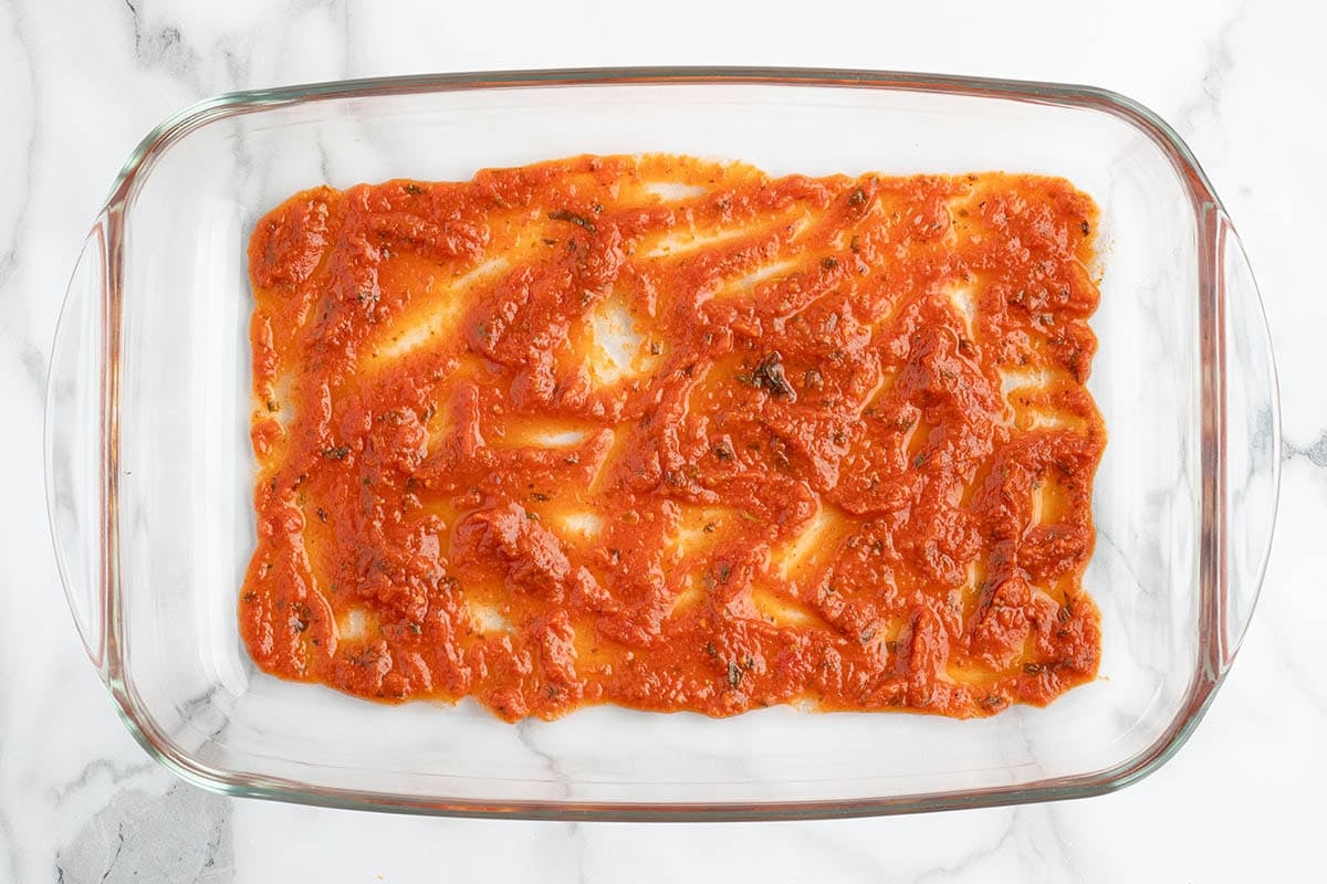 Marinara sauce spread over frozen ravioli in a glass baking dish.