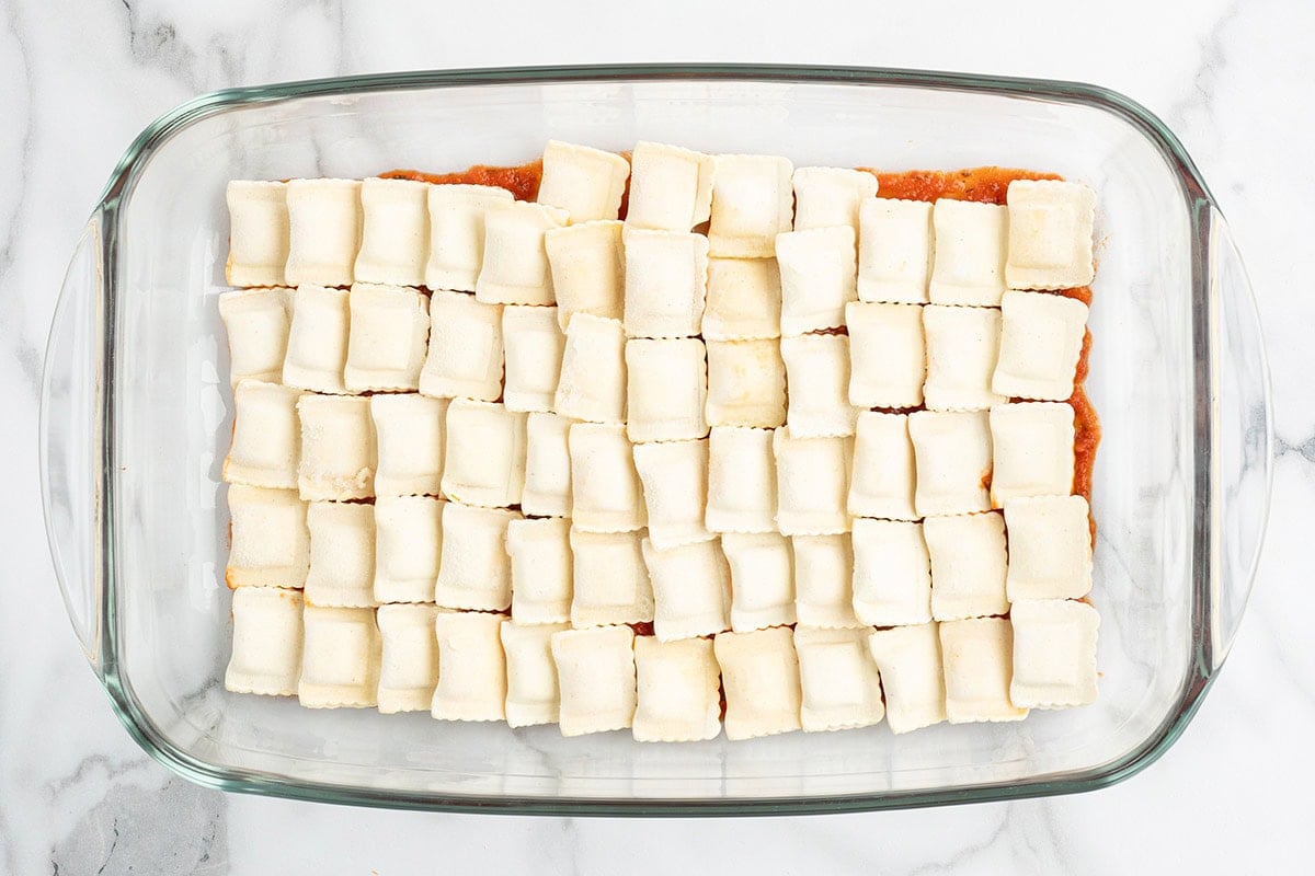 Frozen ravioli arranged in rows over marinara sauce in a baking dish.