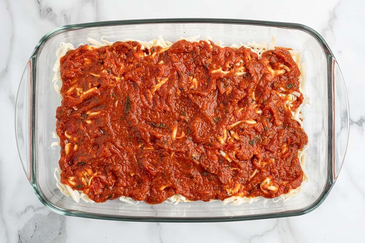 Layer of marinara sauce spread over shredded cheese in a ravioli lasagna dish.