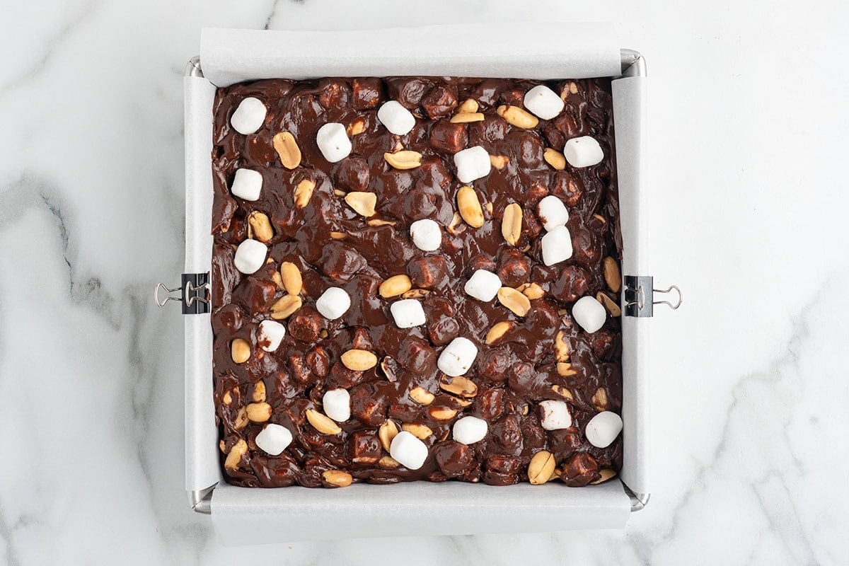 Rocky Road Fudge mixture spread into a parchment-lined pan and topped with extra mini marshmallows and crunchy peanuts before chilling.