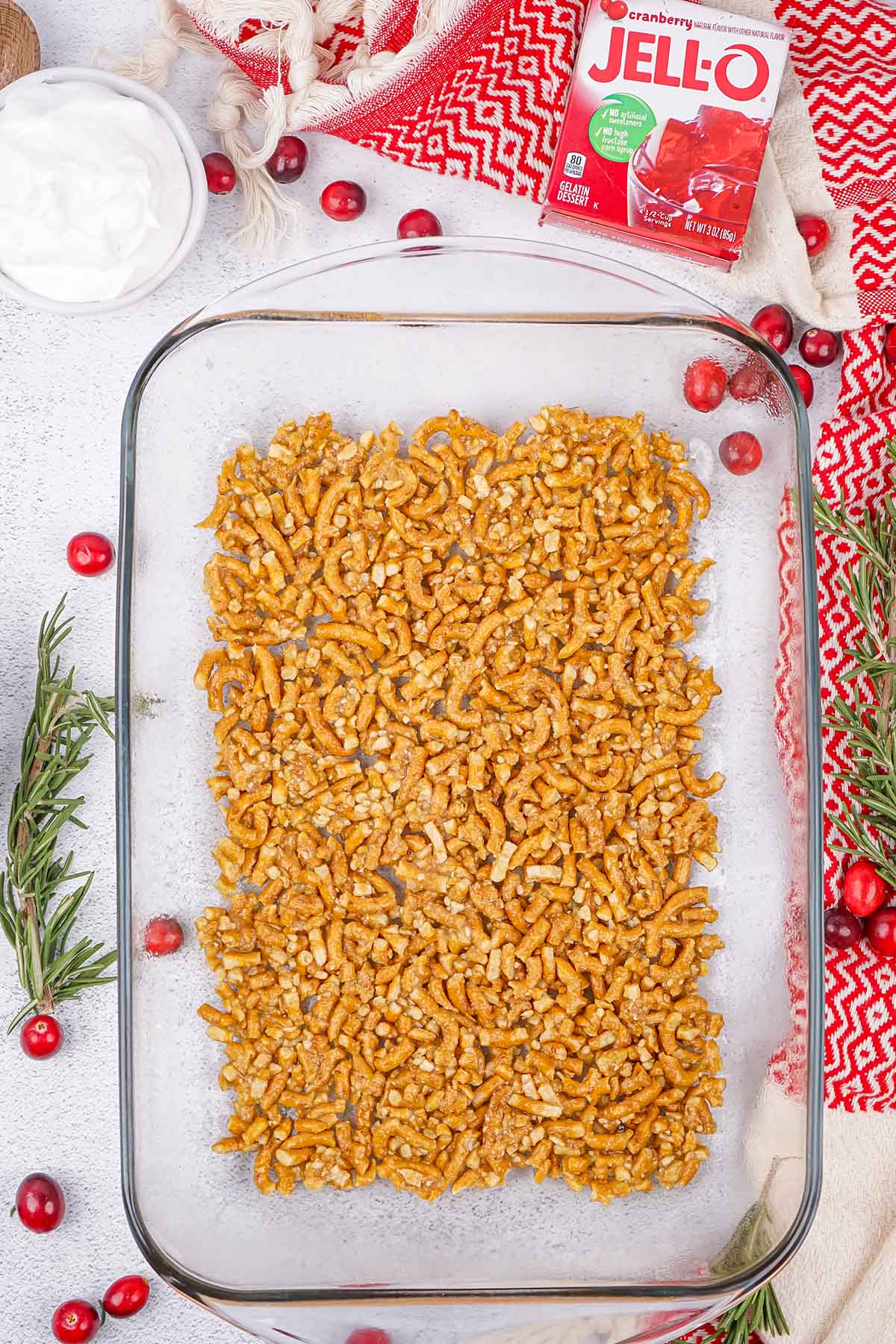 Crushed pretzel crust pressed into a glass baking dish, surrounded by cranberries and rosemary.