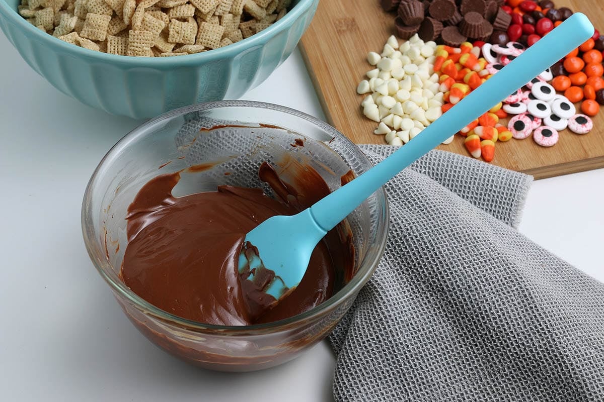 Bowl of melted chocolate with a blue spatula, next to Halloween candies and cereal ready for Halloween puppy chow.