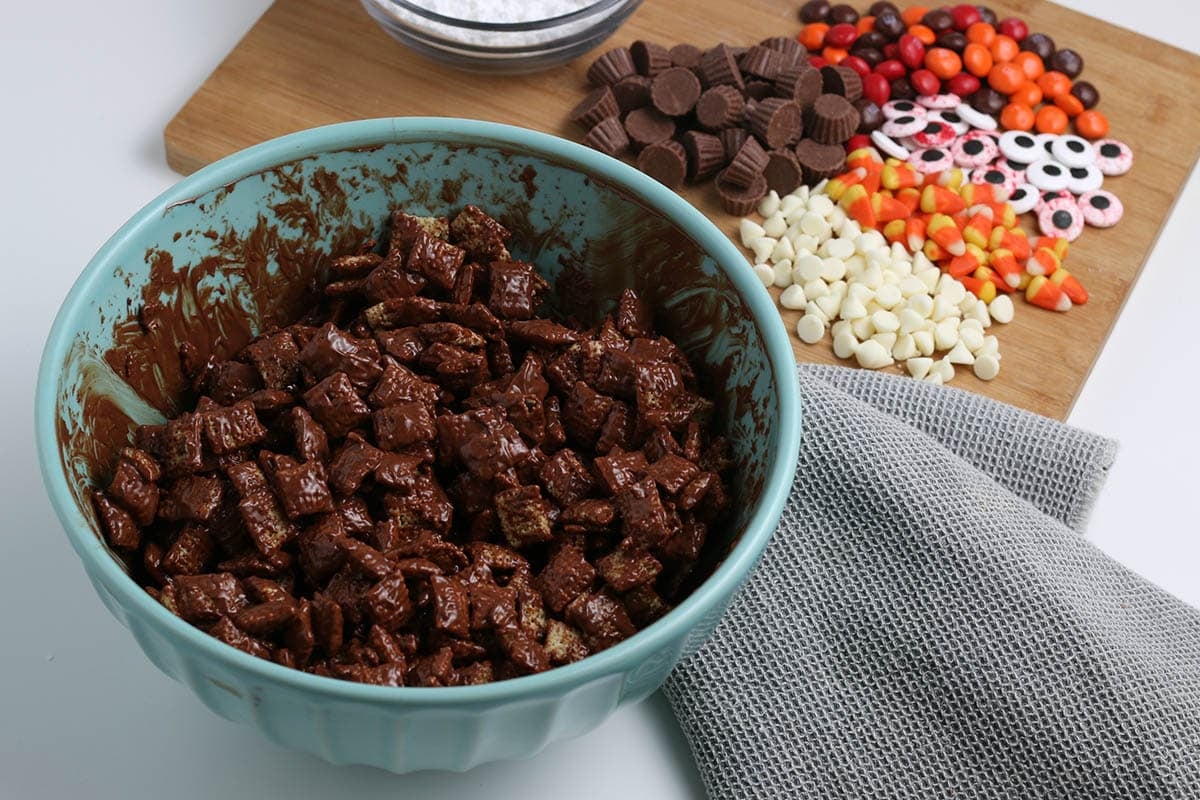 Chex cereal coated in melted chocolate in a blue bowl, with Halloween candy mix-ins on a cutting board.