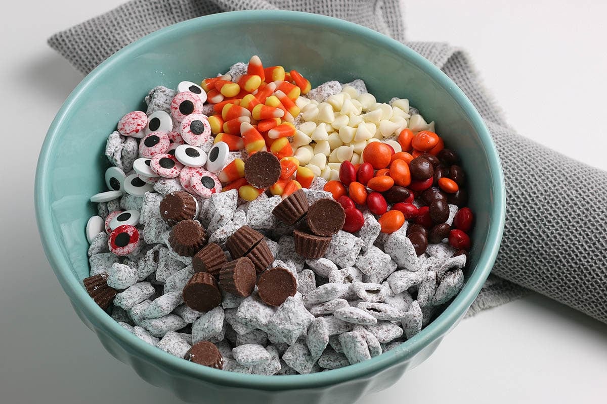 Halloween puppy chow in a blue bowl topped with candy eyeballs, candy corn, mini peanut butter cups, and chocolate candies.