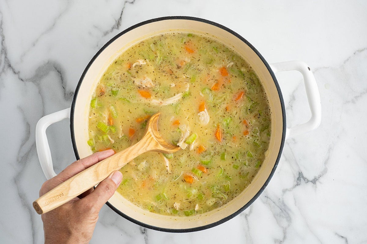 Hand stirring chicken soup with carrots and celery in a pot.