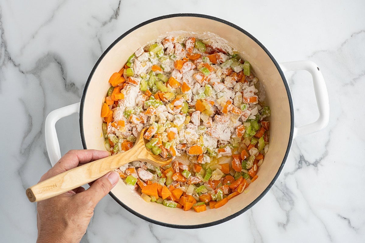 Hand stirring flour into diced vegetables and chicken in a pot.