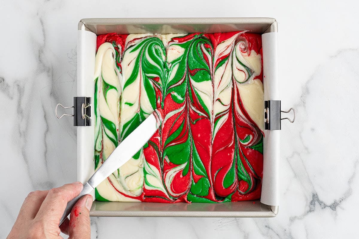 Swirling red, green, and white fudge in a lined square pan with a knife.