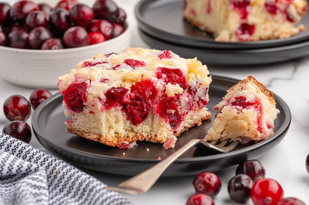 Cranberry cake slice on a black plate with a bite taken on a fork.