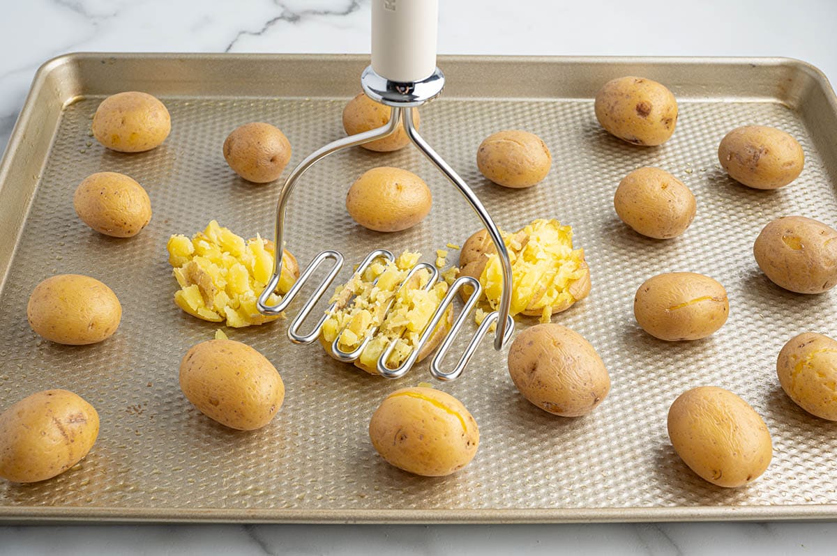 Potato masher smashing boiled potatoes on a baking sheet.