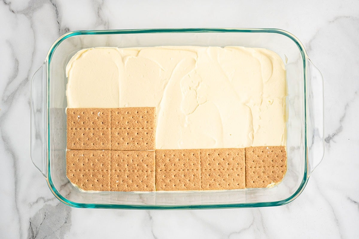 Graham crackers being layered over a pudding filling in a glass baking dish for eclair cake.