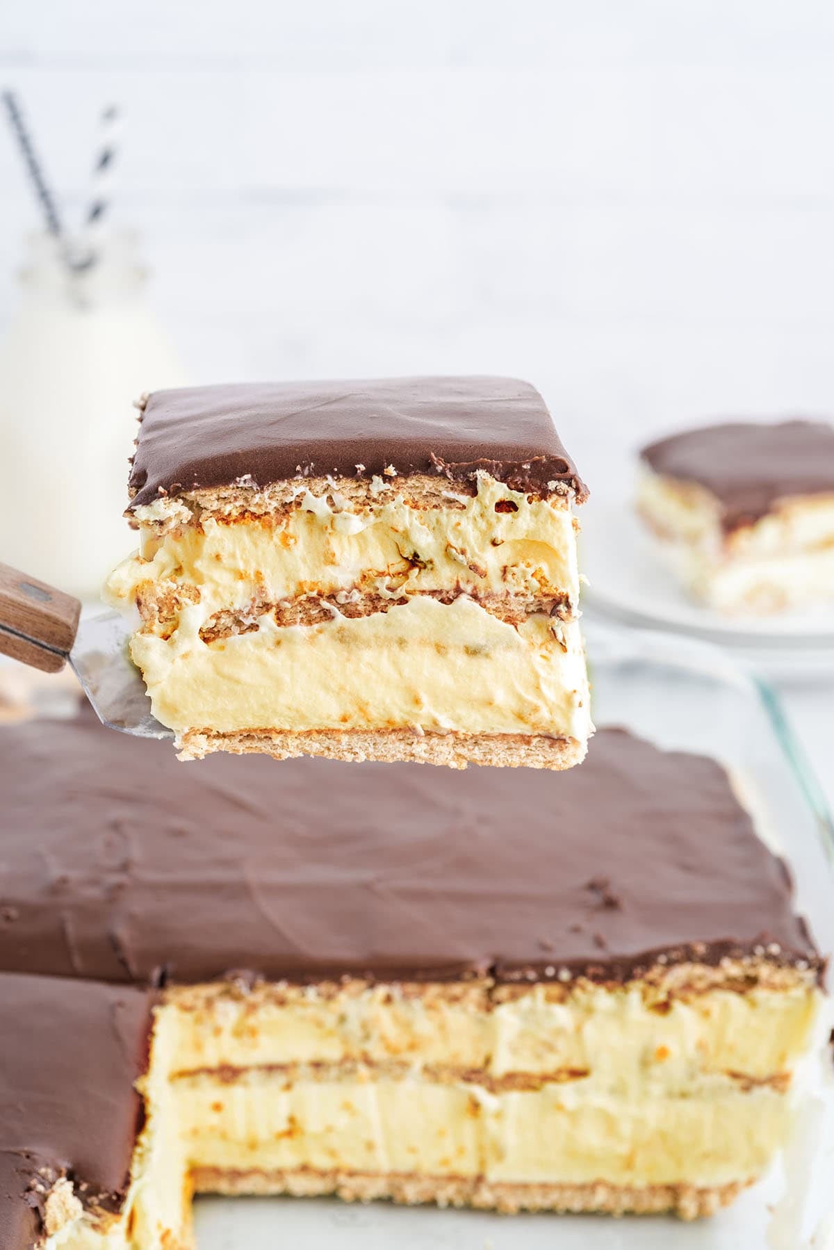 A slice of eclair cake lifted on a server, with the full cake visible in the background.