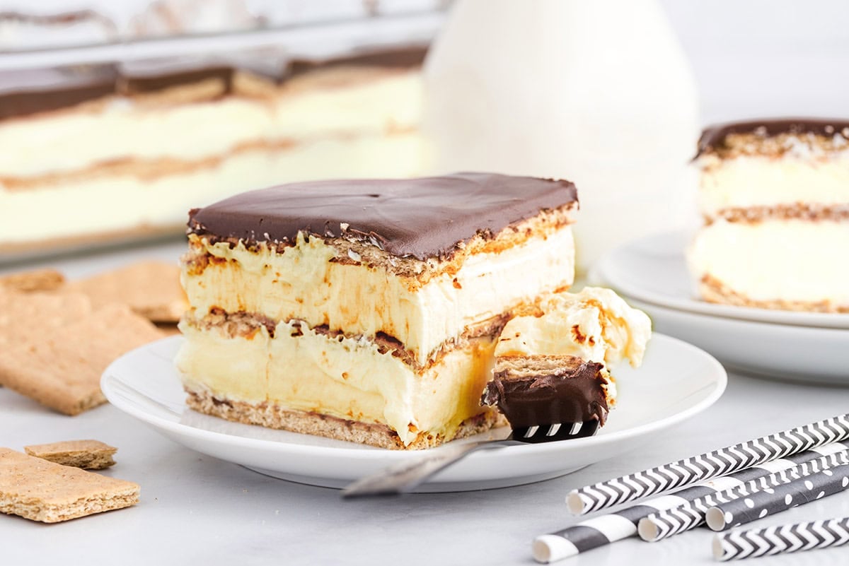 Slice of eclair cake with layered graham crackers, vanilla pudding, and chocolate topping, served with a fork on a plate.