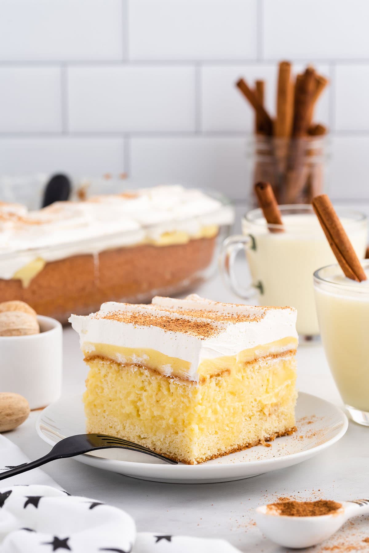 Slice of eggnog poke cake topped with whipped cream and cinnamon, served on a plate with eggnog and cinnamon sticks.