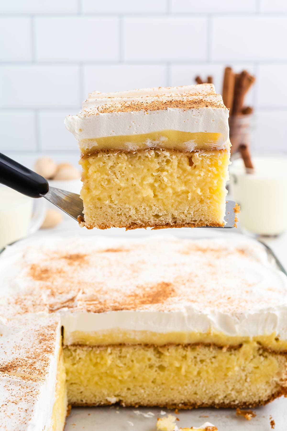 A slice of eggnog poke cake lifted on a spatula, showing its layered interior against a cinnamon stick background.