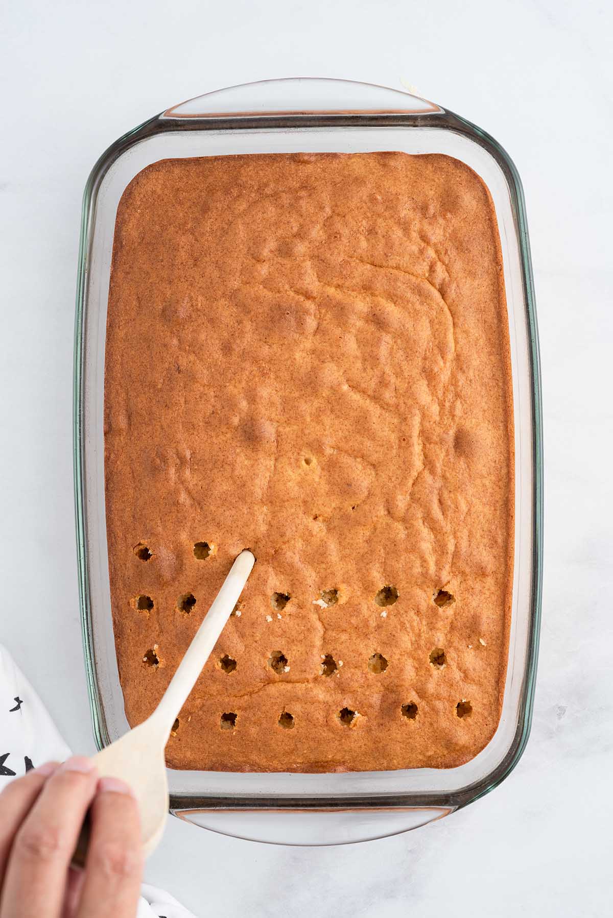 Baked cake in a glass dish being poked with a wooden spoon handle to make holes for the eggnog soak.
