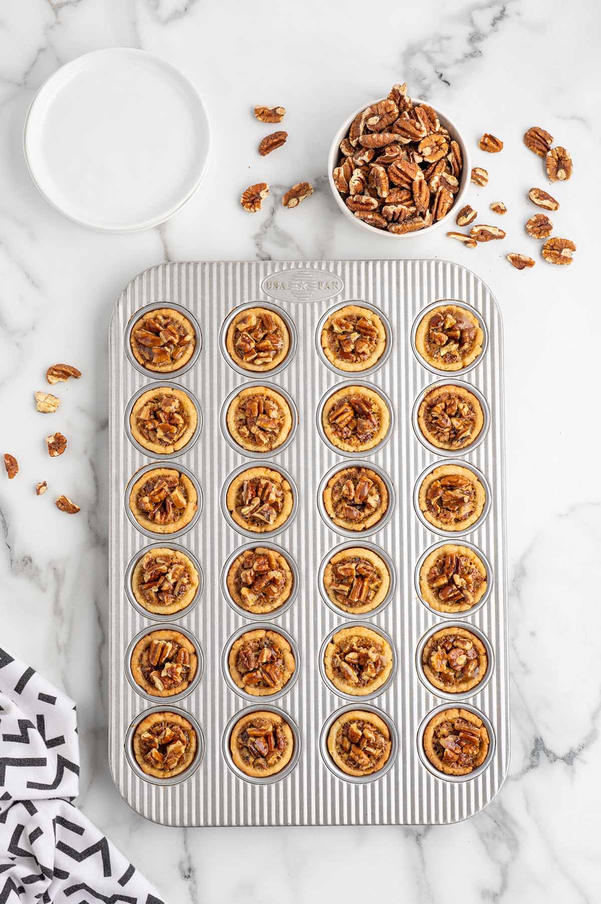 Baked mini pecan pies in a muffin pan with a bowl of pecans and a kitchen towel nearby.