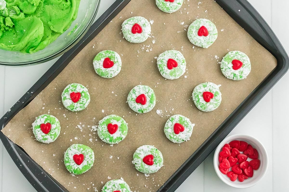 Unbaked Grinch cake mix cookies topped with red candy hearts on a parchment-lined baking sheet.