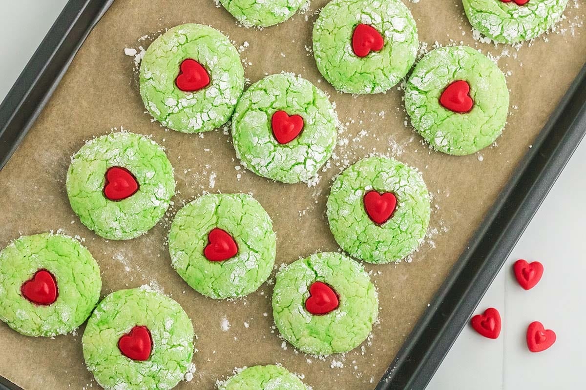 Baked green Grinch cake mix cookies with red candy hearts on a baking sheet.