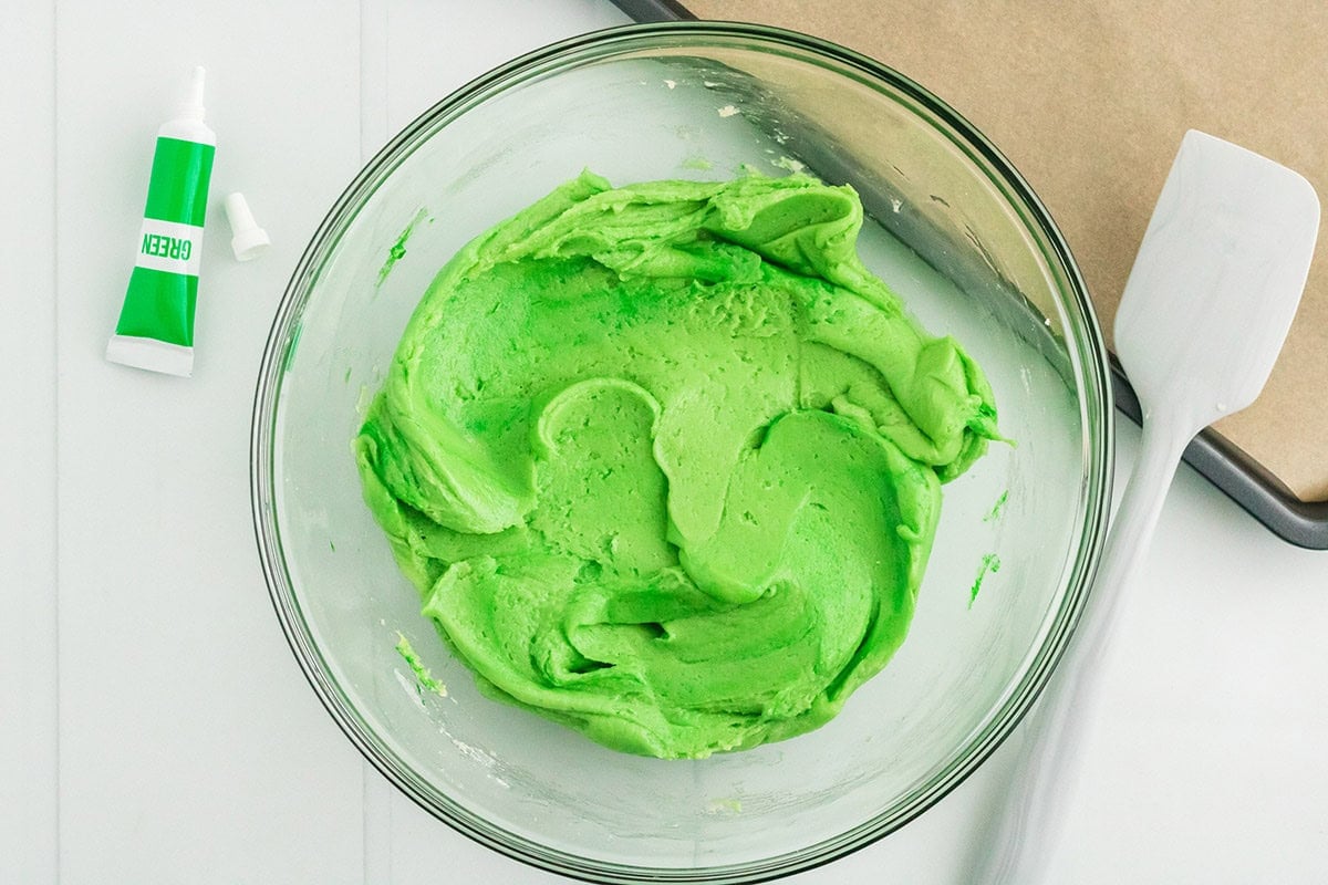 Bright green Grinch cake mix cookie dough in a glass mixing bowl with green food coloring and a spatula.