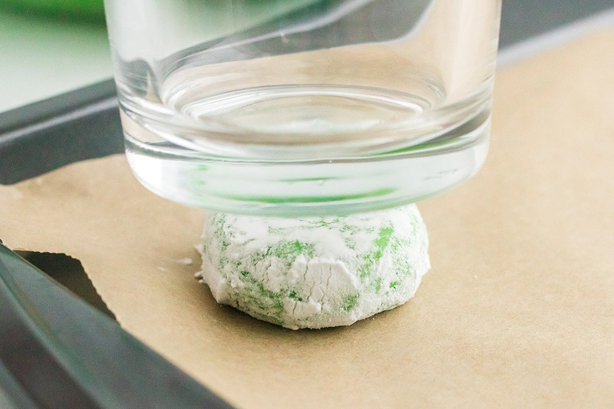Green Grinch cookie dough coated in powdered sugar being flattened with a glass.