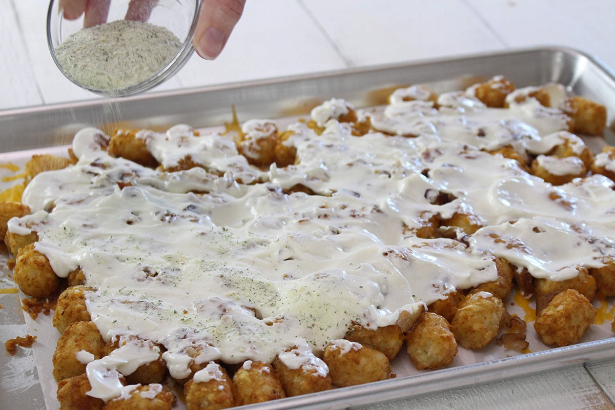 Ranch dressing drizzled over cheesy tater tots, with seasoning being sprinkled on top.