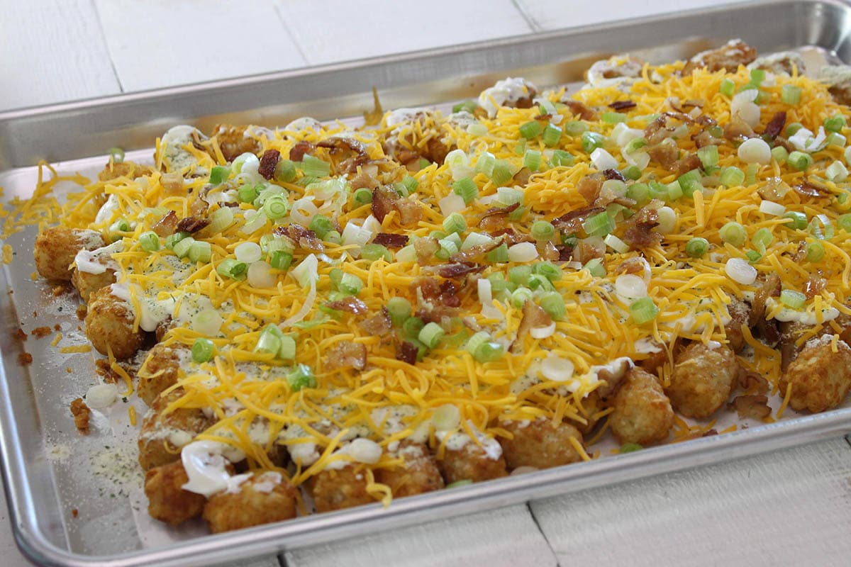Loaded tater tots topped with melted cheese, ranch, bacon bits, and chopped green onions on a baking sheet.