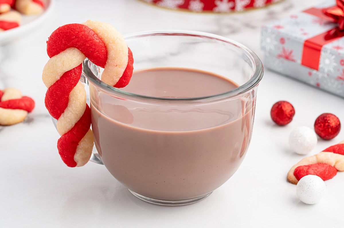 Candy cane cookie hooked on the rim of a glass mug filled with hot chocolate.
