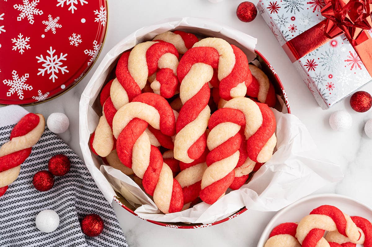 Candy cane-shaped cookies piled in a red snowflake tin lined with parchment paper.