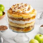 Caramel apple trifle with layers of cake, pudding, whipped cream, and pecans, topped with caramel apples, whipped cream, and caramel drizzle.