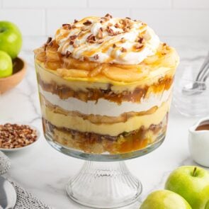 Caramel apple trifle with layers of cake, pudding, whipped cream, and pecans, topped with caramel apples, whipped cream, and caramel drizzle.