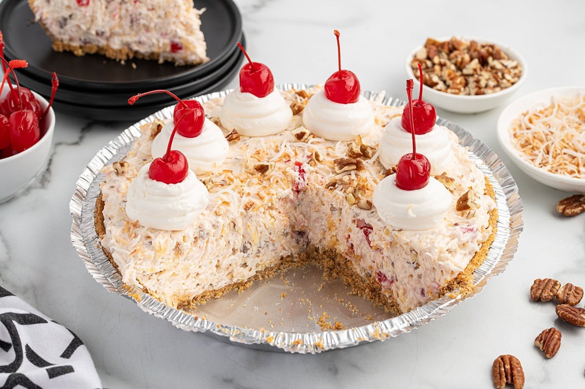 Millionaire pie with a slice removed, topped with whipped cream, cherries, coconut, and pecans.