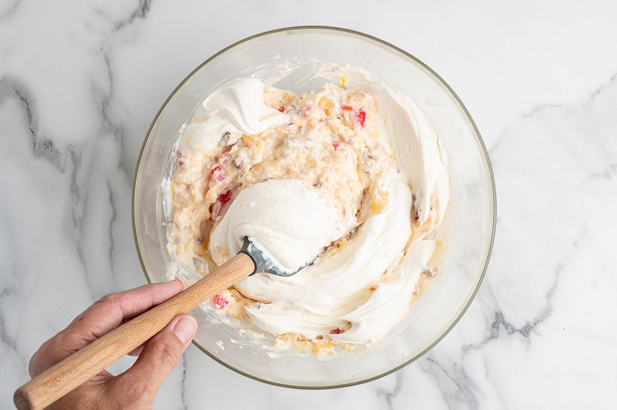 Whipped cream being folded into millionaire pie filling with fruit and nuts.