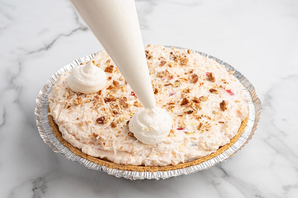 Whipped cream being piped onto millionaire pie topped with coconut and pecans.