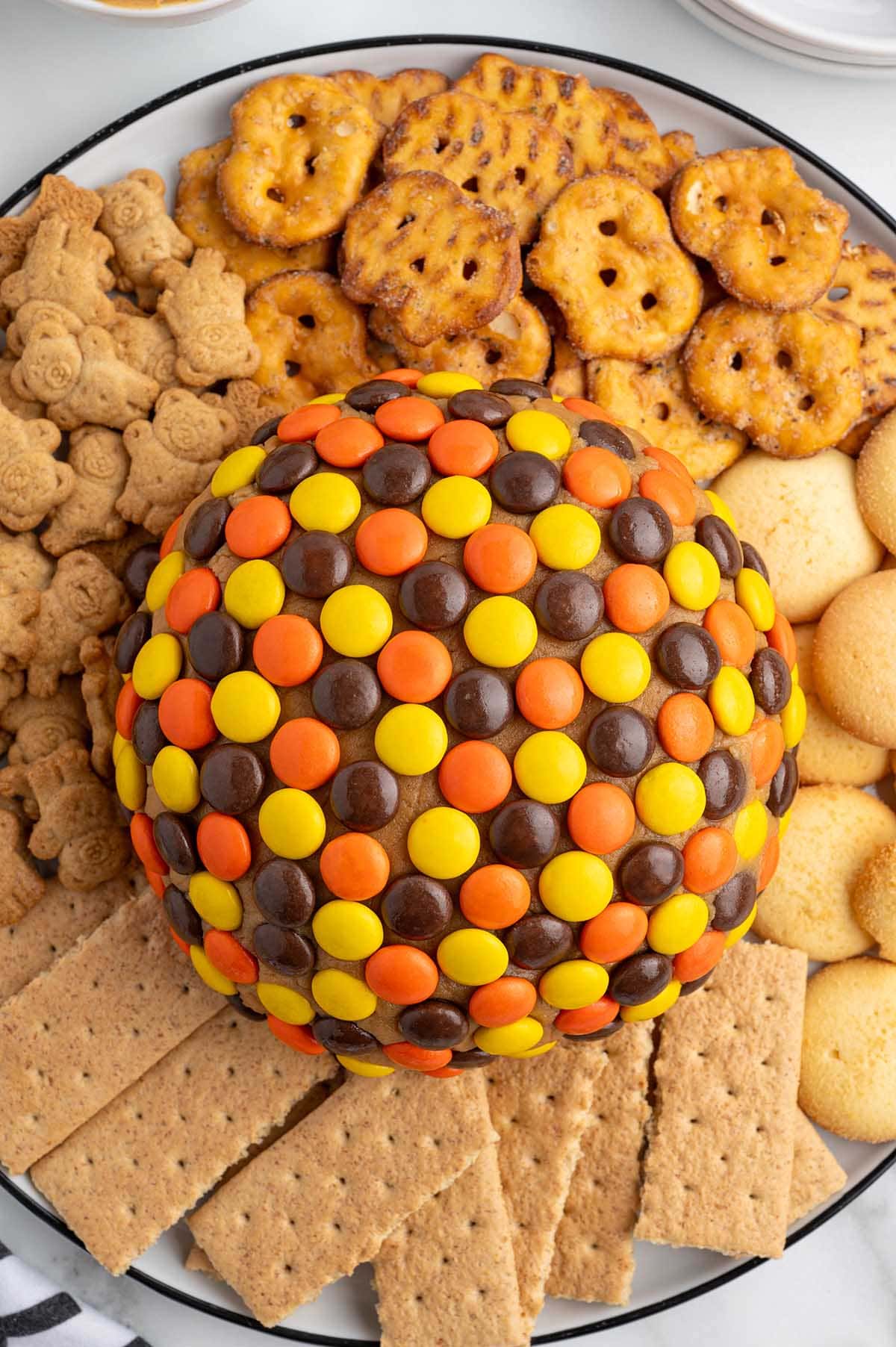 Overhead view of a peanut butter cheese ball covered in Reese's Pieces, surrounded by pretzels, graham crackers, animal crackers, and vanilla wafers.