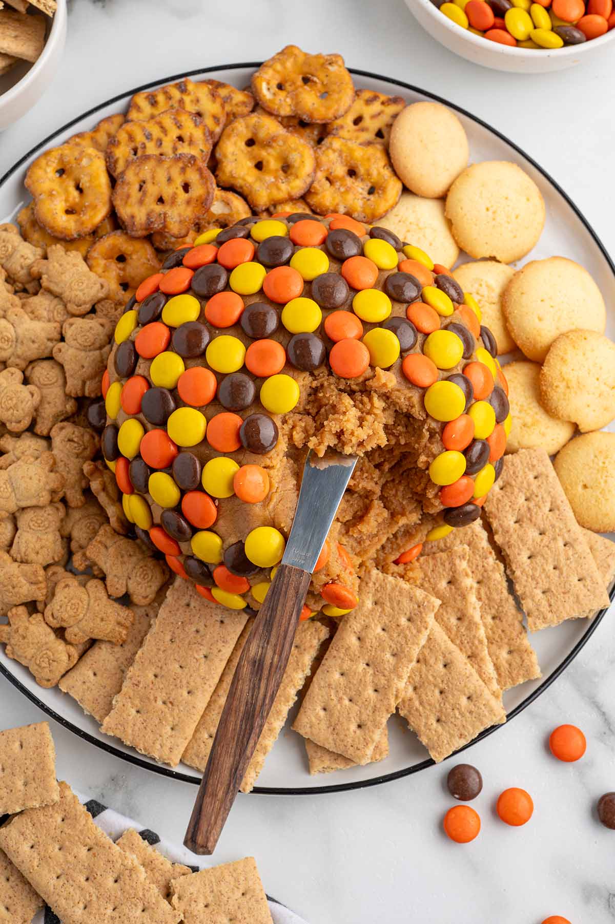 Knife scooping into a peanut butter cheese ball covered in Reese's Pieces, surrounded by crackers, pretzels, and cookies.