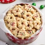 Tin of spritz cookies shaped like trees and wreaths with sprinkles, lined with parchment.