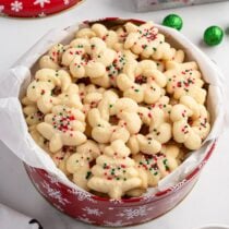 Tin of spritz cookies shaped like trees and wreaths with sprinkles, lined with parchment.