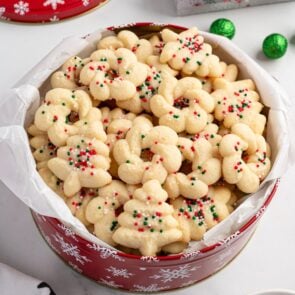 Tin of spritz cookies shaped like trees and wreaths with sprinkles, lined with parchment.