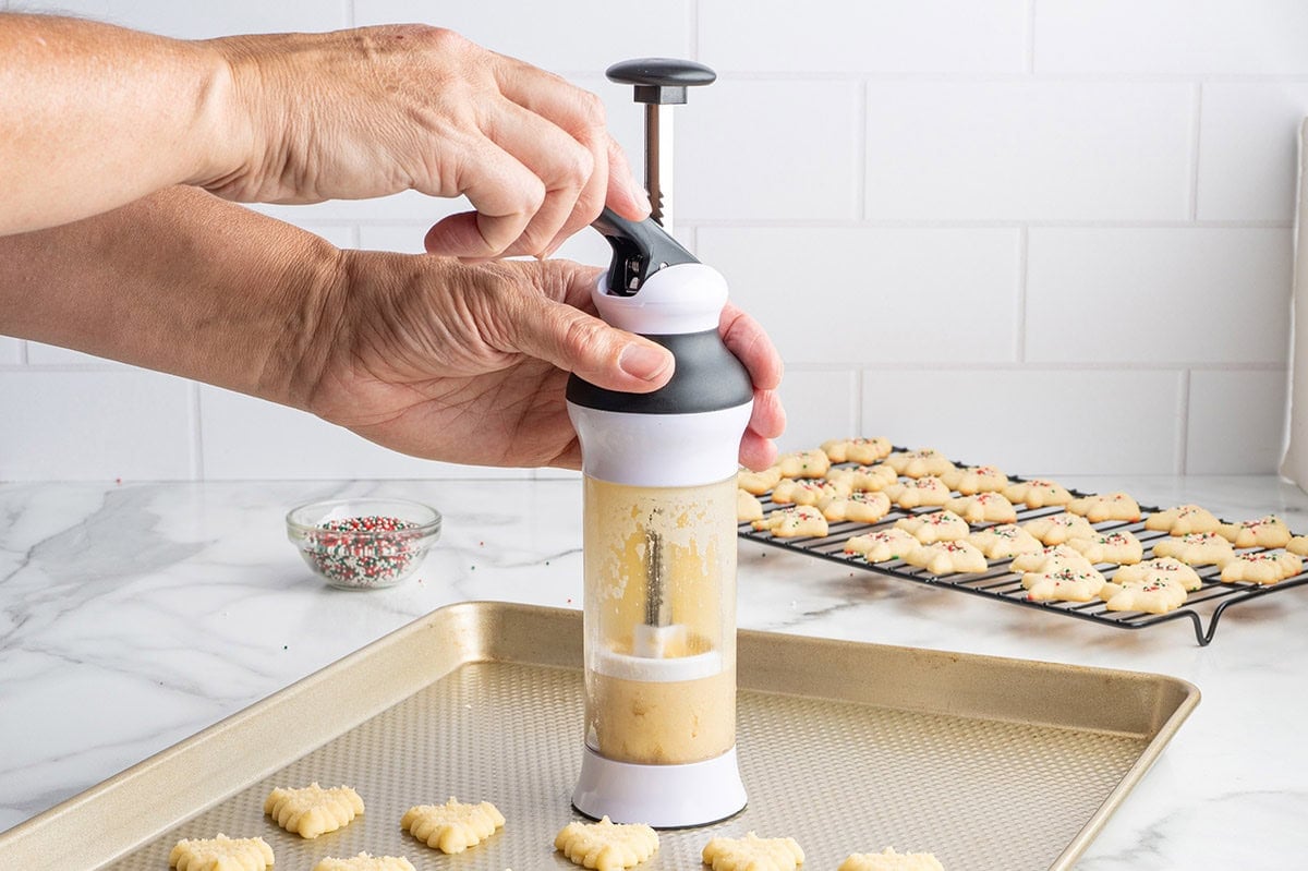 Hands pressing spritz cookie dough from a cookie press onto a baking sheet.