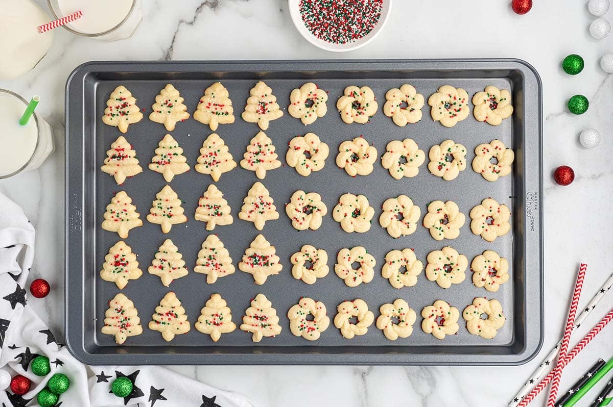 Baked tree and wreath spritz cookies with sprinkles on a baking sheet.
